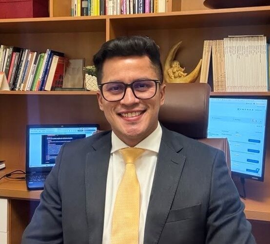 Dr. Williams Silva sorrindo em seu escritório, de terno e gravata, diante da estante de livros